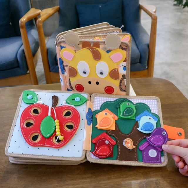 Photographie d'un livre Montessori en tissu, avec une couverture en forme de tête de girafe, exposé sur une table. Le livre est ouvert sur deux pages interactives : l'une montre une pomme rouge avec des trous, idéale pour des activités de motricité fine, et l'autre une scène d'arbre avec des oiseaux colorés dans leurs nids.