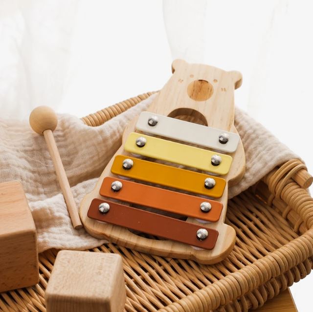 Xylophone bébé en bois en forme d'ours composé de lames colorées, accompagné d'un maillet en bois, posé dans un panier en osier avec des cubes en bois.