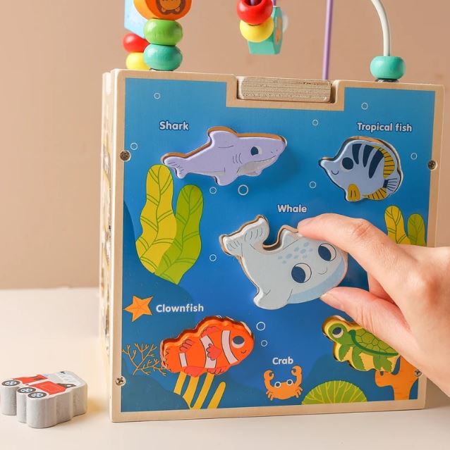 Enfant jouant avec un cube en bois multi activités, insérant des pièces en forme d'animaux marins comme la baleine, le poisson clown et le requin pour favoriser l'apprentissage et la motricité.