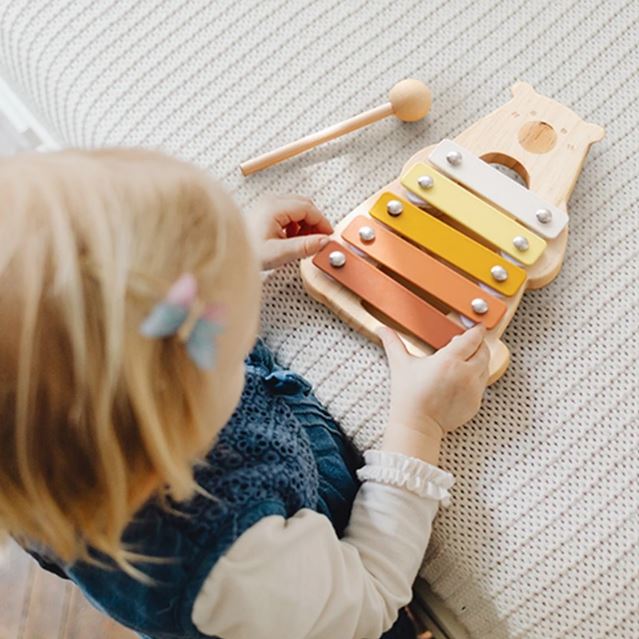 Un enfant jouant avec un xylophone en bois pour bébé en forme d'ours, aux lames colorées, posé sur un tapis, avec un maillet en bois à côté.